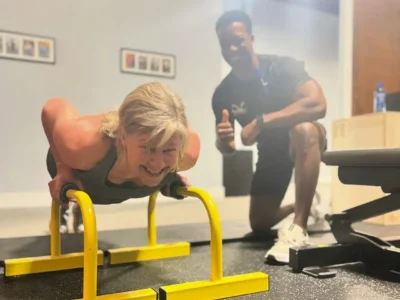 Woman performing push-ups on parallel bars while a trainer encourages her